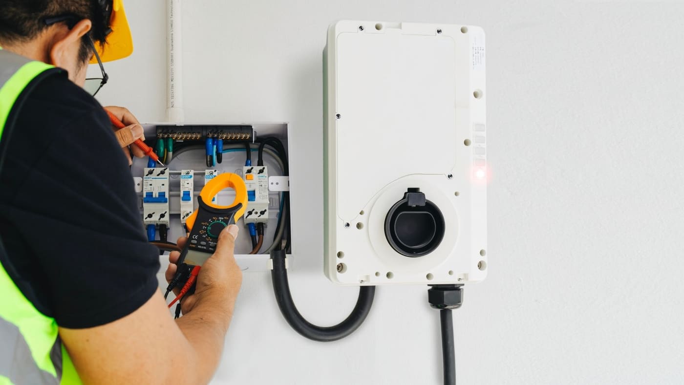 Electrician testing a garage subpanel beside a wall-mounted EV charging unit